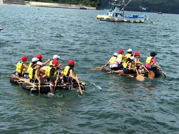 黄色のライフジャケットを着用した子供たちが手に木製のパドルを持ち水をかきながら一生懸命に前へ進もうとしている2台の筏に乗った子どもたちのグループを写した海上の写真