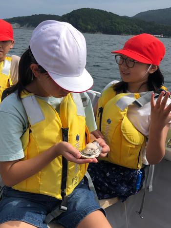 黄色いライフジャケットを着用し海の上に浮かぶ船の上で白い帽子をかぶった女の子が手のひらに小さな貝のようなものを乗せてじっと見つめその隣の赤い帽子と眼鏡をかけた女の子が少し驚いた表情で手を広げて話している写真