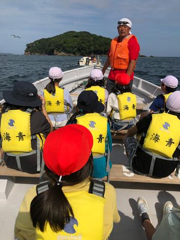 小型ボートの上で赤い救命胴衣を着た男性指導者が立って黄色いライフジャケットを身につけた子どもたちが正面を向いて男性の話を真剣に聞いている写真