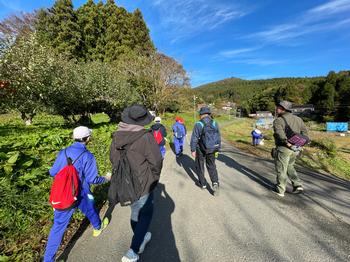 晴天の中ジャージ姿の子供たちが地域の人と道路を歩いている様子を後方から写した写真
