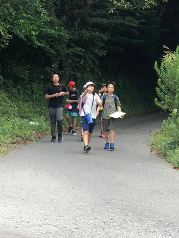 緑の中、水筒と地図のようなものを持ちながら前方を指差しして歩いている生徒たちの写真