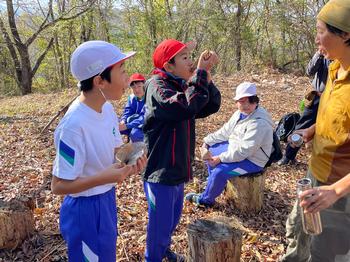 丸太の椅子で休憩している子供と大きな口でピザを口に入れる男子児童、周りで見ている男子児童と女性の写真