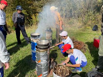ドラム缶の上に鍋を置き、児童がドラム缶に薪をいれ、けむりが出ている様子を写した写真