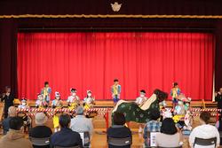 舞台の上で和太鼓を叩く子どもたちと獅子舞が登場し、観客が見守る中で郷土芸能を披露している写真