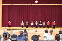 赤いカーテンを背景に、カラフルな服を着た子どもたち8人が舞台の上に並んで立ち、観客が椅子に座って見守っている写真