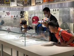 帽子をかぶり、マスクをつけ、リュックを背負った数人の児童たちが、ガラスケースの中の展示物を見ている写真