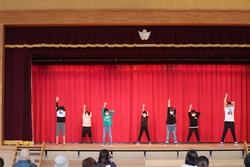 赤い幕の前の舞台で、子どもたち7名が一列に並んで右手を高く上げてポーズをとっている写真