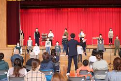 赤い幕の前の舞台で子どもたちが立って合唱し、観客席には多くの人が座って見守っている写真