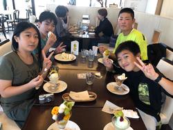 屋内のカフェで5人の児童たちがテーブルを囲み、それぞれパフェやアイスクリームのようなスイーツを食べ、カメラに向かってピースサインをしている楽しげな様子を写した写真