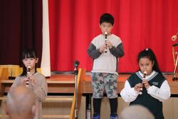 赤い幕の前の舞台で、3人の子どもたちがリコーダーを演奏している写真