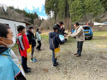 晴れた日の屋外で、黄色い肩掛けカバンを下げた子どもたちが集まり、ひとりの大人が代表の子どもに何かを渡している光景の写真