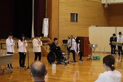 車椅子の人を含むグループがマイクを使って楽器演奏や発表をしている様子の写真