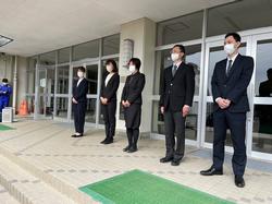 校舎の入り口前にスーツを着た男性2名、女性3名の職員が横一列に並んで立っている写真