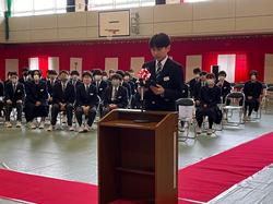 紅白幕が張られた体育館に並んだ椅子に生徒たちが座り、その前にある演台のマイクに向かって男子生徒が原稿を読んでいる様子の写真