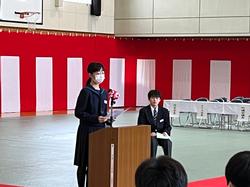 紅白幕が張られた体育館で、演台に立ち、マイクに向かって話をする女子生徒の写真