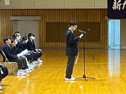 椅子に座る生徒たちを背後に、スタンドマイクの前に立った男子生徒が原稿を読んでいる写真