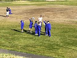 芝生の運動場に集まった、ジャージ姿の10人ほどの生徒がカメラに手を振っている写真