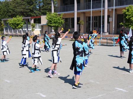 校舎前のグラウンドで、白と水色のお揃いの法被姿の大勢の子供たちが両手に持った鳴子を振り上げて踊っている様子を右後方から撮影した写真