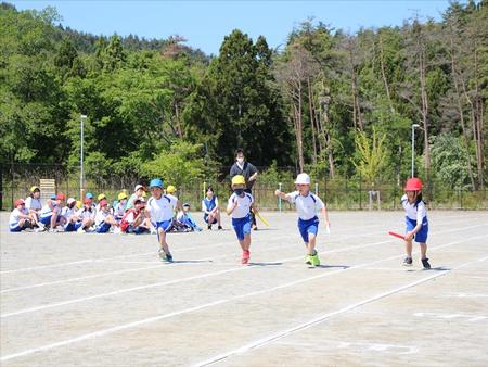 木々に囲まれた晴れた運動場で、トラックの外側に立っている男性教師や、体操服姿の他の児童たちが座って見守っている中、カラフルな色の帽子を被った4人の児童たちがバトンを持って走りだしたリレーの練習の様子写真