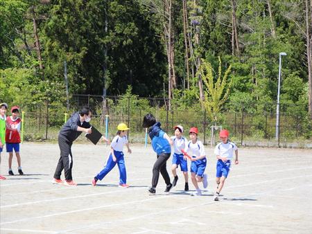 校庭で他の児童たちが見守る中、女性教師や子供たちがバトンを渡したり、受け取ったりしながら走っているリレーの練習を行っている写真