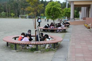 学校の校庭に設置された丸いベンチの中で子どもたちが座って草取り作業をしている様子の写真