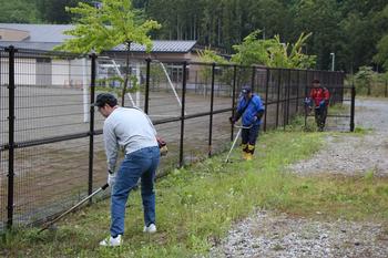3名の作業者が黒いフェンスの近くを草刈り機で草刈りをする様子の写真