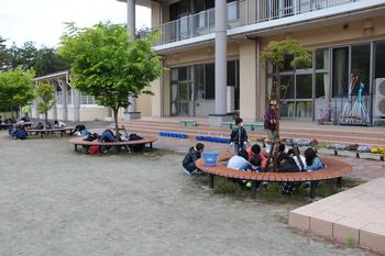 学校の校庭に設置された丸いベンチの周りで、子どもたちが座って草取り作業をしている様子の写真