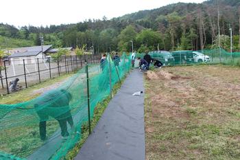 奥に保護者の方が集まり、緑の網の手前に防草シートが貼られた写真