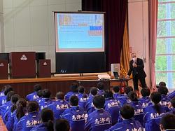 ステージに置かれた大きなスクリーンに資料が映し出され、整列して座った生徒たちがそちらを見る中、前に立った男性がマイクで話をしている写真