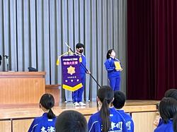 生徒たちが整列する中、ステージの上に旗を持った男子生徒と表彰状を持った女子生徒が立っている写真