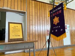 机の上に額に入った表彰状が立てられ、横に旗が並べて置かれている写真