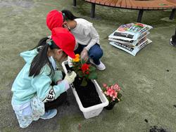 女の子が両手で丁寧に苗を持ち、土の中に優しく植え込んでいるのを横の女の子がサポートしている様子の写真