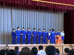 ステージの上に青いジャージを着た生徒たちが横一列に並び、その前でオレンジ色の制服を着た男性が片膝をついている写真
