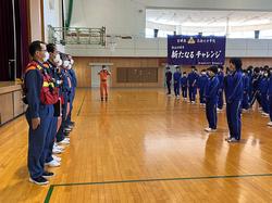 体育館で横一列に並んだ紺色の制服を着た男性たちと向かい合ってジャージを着た生徒たちが整列している写真