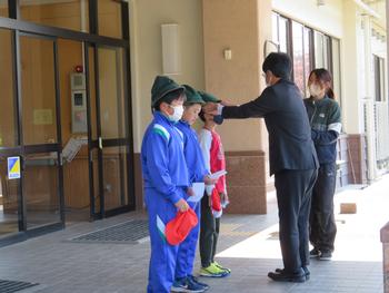 学校の建物の玄関前で、児童3人が並び、正面に立つスーツ姿の男性が真ん中の児童に緑色の帽子を貸与している様子の写真