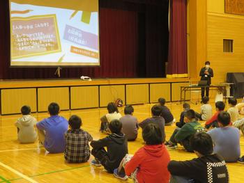 体育館でスクリーンを使った講話を、床に座っている子どもたちが真剣に聞いている様子の写真