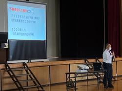 ステージの上にセットされた大きなスクリーンに資料が映し出され、その前に立った男性がマイクで話をしている写真