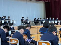 体育館のステージ上に役割の紙が取り付けられた長机が並び、たくさんの生徒たちが座り、ステージ下に並んだ椅子に生徒たちが座っている写真