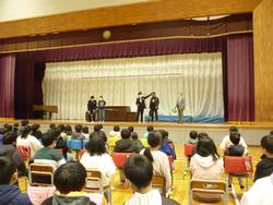 体育館に並べられた椅子に座り体育館の舞台で警察官の方が演技をしているのを見ている児童を後ろから撮影した写真