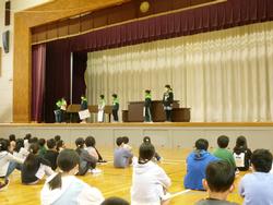 体育館の舞台で肩に黄緑色のバンダナを付けた児童がカードなどを持ち話しているのを床に体育座りをして聞いている児童達の写真