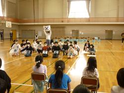 新一年生が座っている前で児童が「おめでとう」とひらがなで書かれた文字を1文字づつ抱え1人の児童が立ってバツと書かれたカードを両手で上げて学校紹介をしている写真