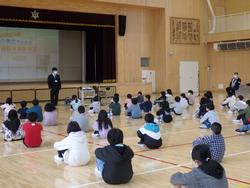 体育館の床に座っている生徒たちがステージ上のスクリーンを見ながら、立って説明する南三陸警察署の方の話を聞いている安全学習の様子の写真