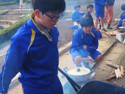 屋外で複数の生徒たちが体操服を着て、薪や大きな鍋など炊き出しの準備をし、1人の男子生徒は一輪車で何かを運んでいる様子の写真