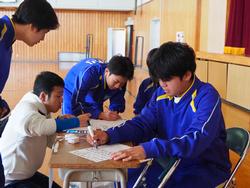 体育館に置かれた机を囲み、体操服を着た複数の生徒たちが用紙に何かを書き込んだり話し合ったりしている様子を写した写真