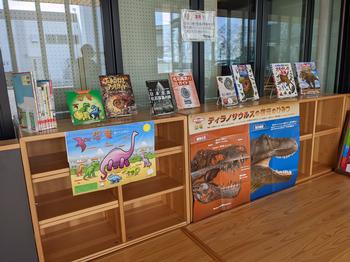 本棚の上段には恐竜に関する本が複数並べられていて、下段には色鮮やかな恐竜のポスターが貼られている図書コーナーの写真