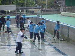 屋外のプールで、複数の生徒たちがほうきやデッキブラシを使ってプールの床を清掃している写真