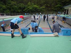 プールの清掃活動で、複数の子供たちがデッキブラシやほうきを使ってプール掃除を行っている写真