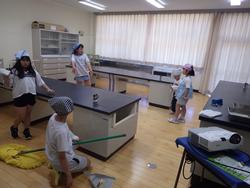 複数の生徒たちが、理科室のような場所で、モップやホウキを使って床を掃除している様子の写真