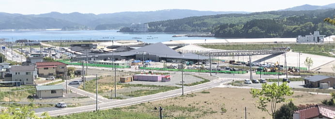 海が広がる、その先には山々が連なり中央には、三角形の屋根が特徴的な大きな建物があり新しい道路や電柱が整備され、まばらに建物が建っている写真