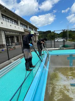 プールサイドに立った男性2名が長靴をはきながらプールの縁や壁に水をかけ掃除をしている写真